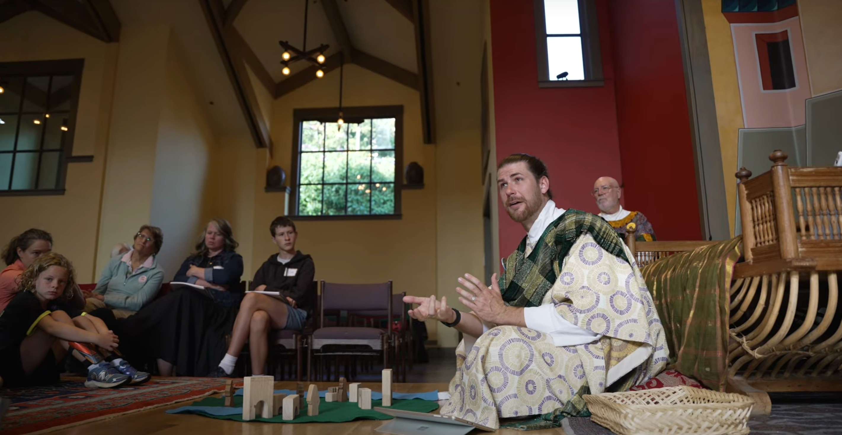 Peter Levenstrong seated on the floor of a church, leading a story with small wooden figures while people of all ages look on