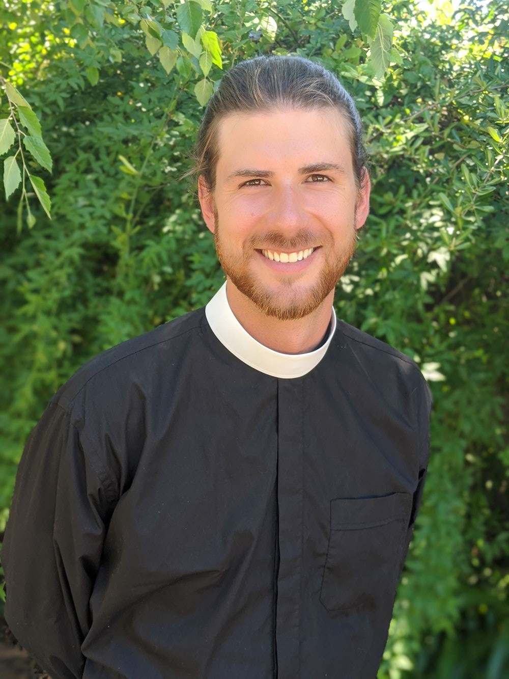 Peter Levenstrong smiling, wearing a black clergy shirt and white collar, in front of green leaves